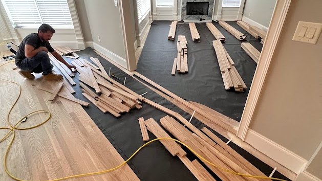 A Hardwood Guys installer laying new hardwood board by board over underlayment, with racked planks spread across the room ahead of him