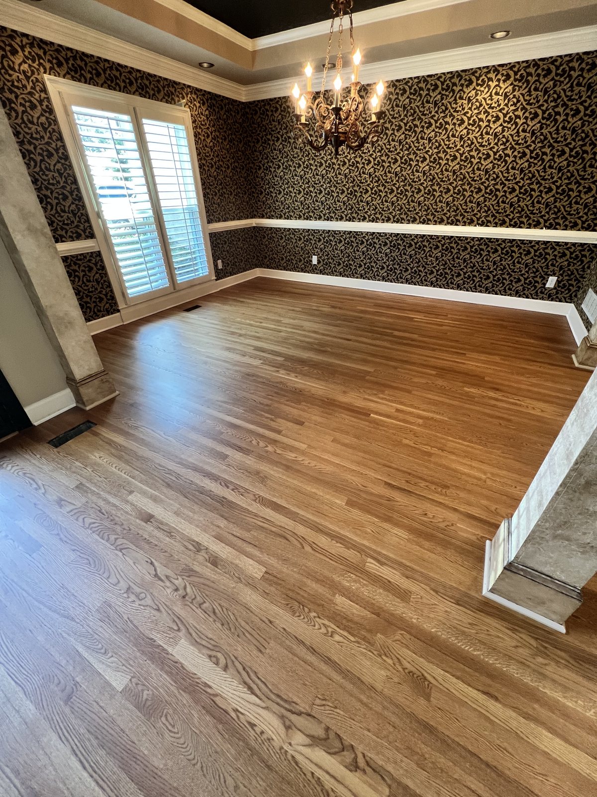 Dining room with red oak hardwood floor in Golden Oak stain and matte finish, patterned wallpaper, chandelier and arched shuttered window