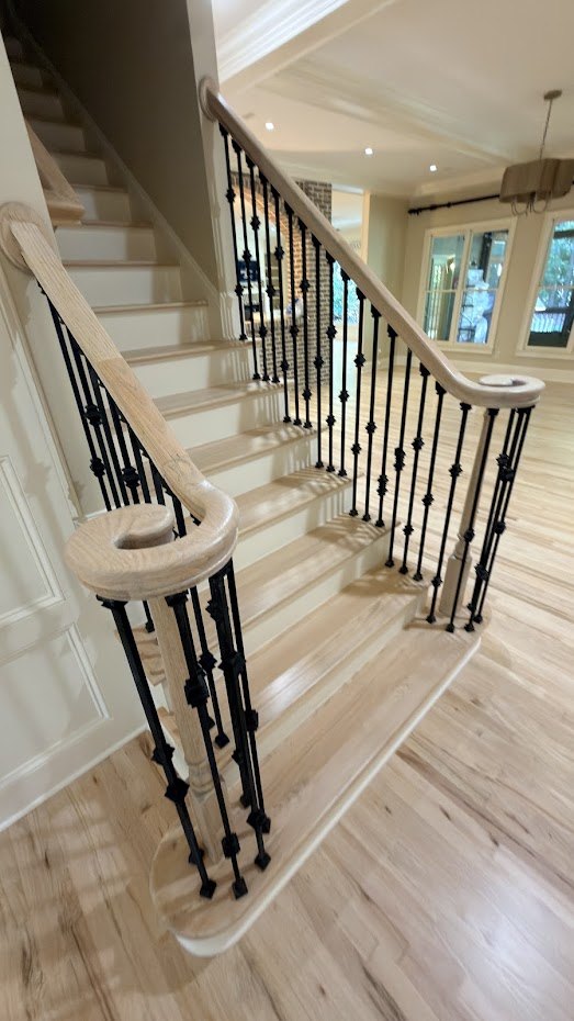 A staircase with light natural-colored oak treads, a curved handrail and newel, and black iron balusters with knuckle details, above a light hardwood floor.